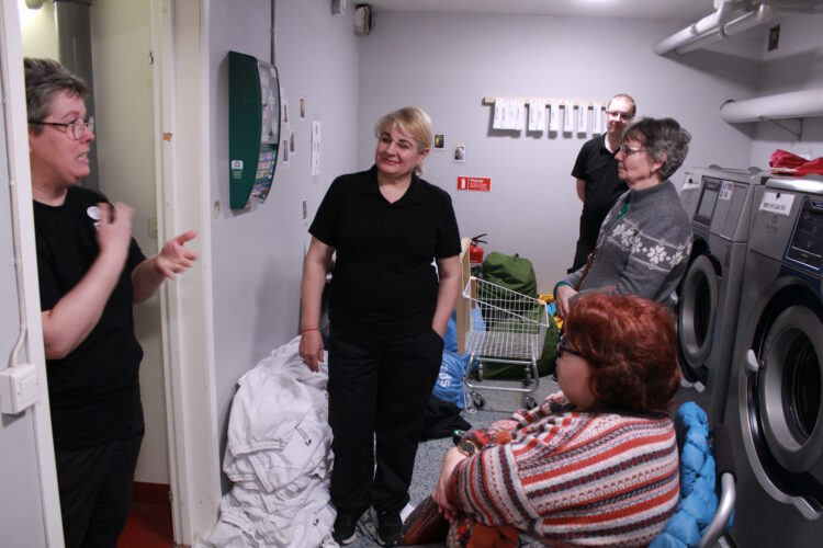 Projektet på Tvätten i Härnösand. Vi är samlade framför en rad med tvättmaskiner. Högar med tvätt på golvet och en tom tvättkorg på hjul. Anita pratar och alla lyssnar på henne.
