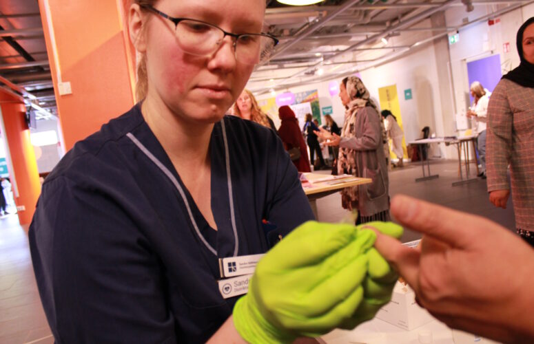 Sjuksyrra med gröna gummihandskar håller i en hand som hon tar blodprov på. Besökare på Welcome-house-event i bakgrunden.