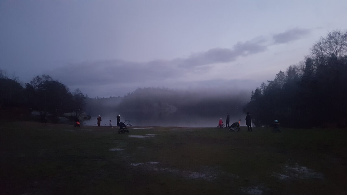 Skymning över Söderbysjön. Marken i förgrunden nästan svart, sjön som en ljusare grå strimma, svarta trän på andra sidan sjön och över alltihop en himmel i många olika grå toner. Människor i siluett framför sjön.