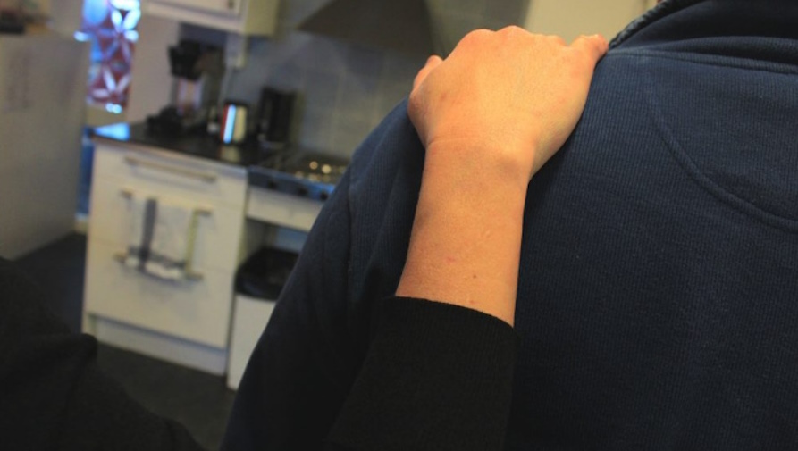 En persons hand på en annans axel mot oskärpt köksbakgrund.