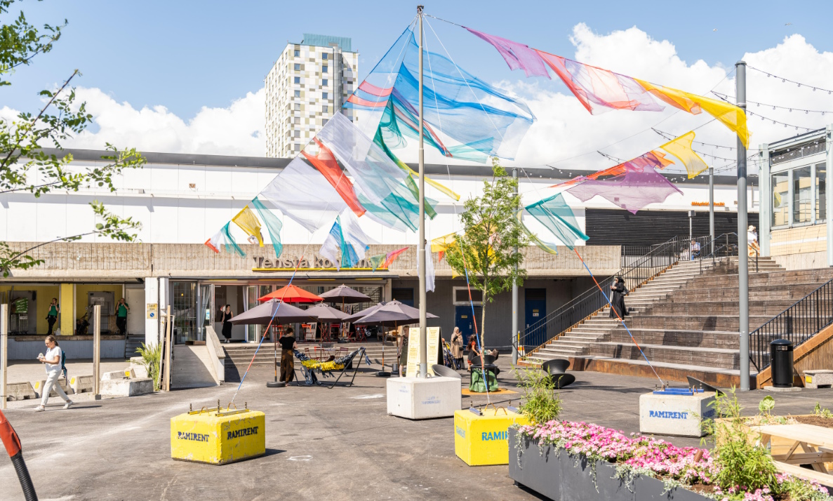 Förortstorg en blåsig sommardag med vimplar i olika färger i luften. Ingången till Tensta konsthall och blå himmel med cumulusmoln.