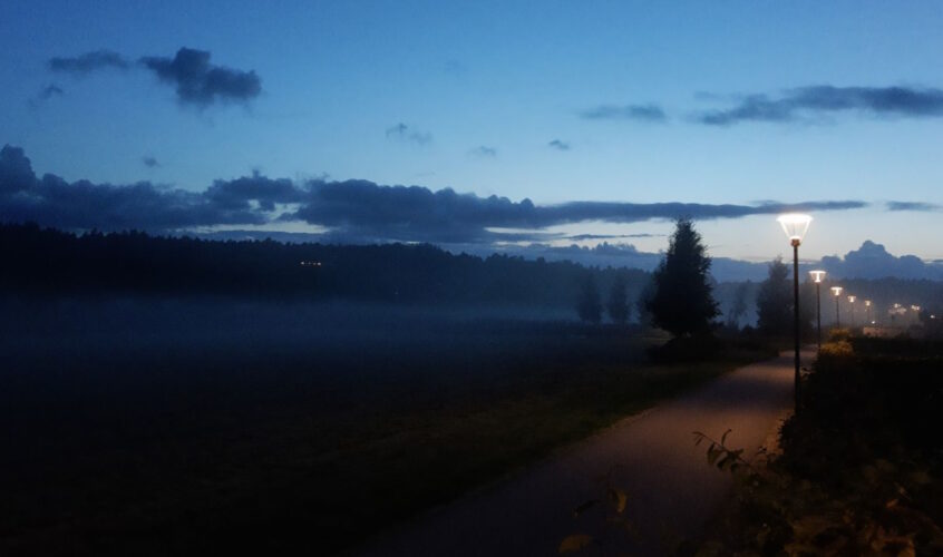 Solen har just gått ner och lyser upp himlen i olika blå nyanser. Till höger ett promenadstråk med gula lampor och till vänster skog i mörker. Höstkänslor.