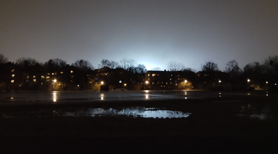 Bild tagen kvällstid. Mörker i förgrunden och himlen upplyst av stadens ljus. Däremellan bostadshus med jlus i fönstren och framför det en stor vattensamling som speglar ljuset från himlen.